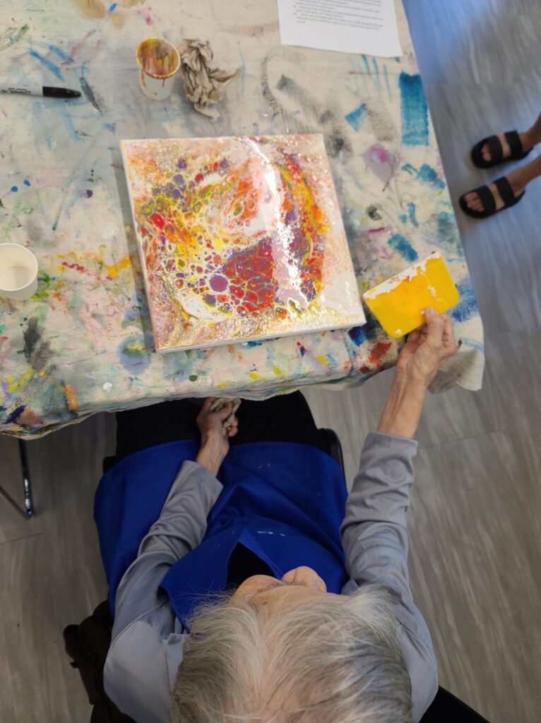 A 96-year-old woman viewed from above creating a colorful fluid acrylic painting using a swipe technique in a library art class.