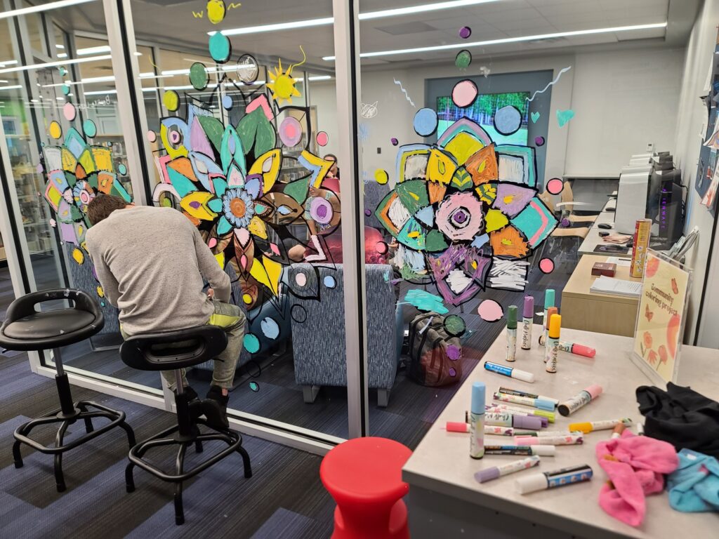 A person seated at a stool coloring a glass mandala while art markers and supplies sit on a nearby table.
