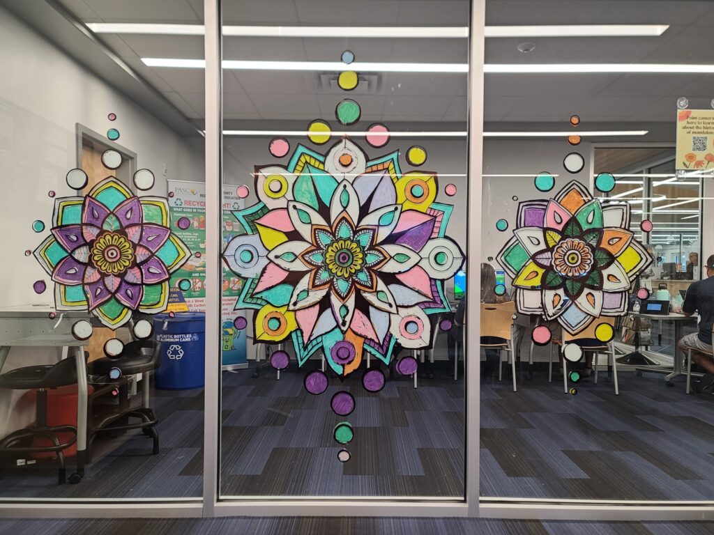 Three finished window mandalas displayed on large glass panels in the teen room, viewed from inside.
