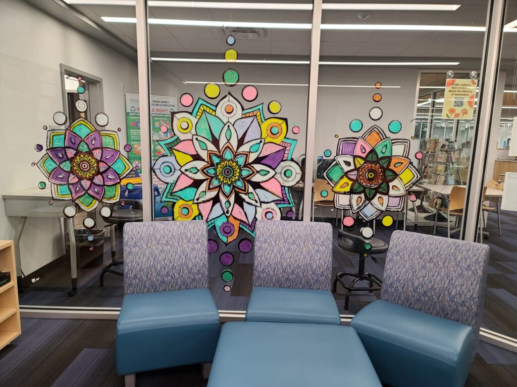  Three finished window mandalas displayed on large glass panels in the teen room, viewed from inside with blue seating in front.