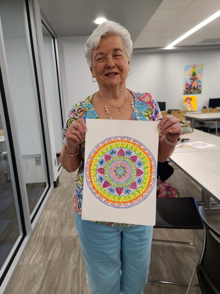 An older woman smiles and holds up a brightly colored geometric mandala she completed in a drawing class.
