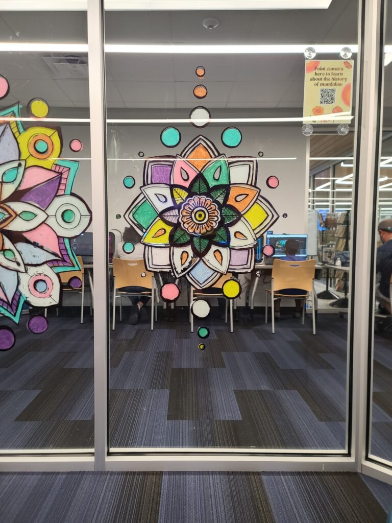A brightly colored mandala with teal, green, yellow, pink, and purple shapes painted on a glass panel.

