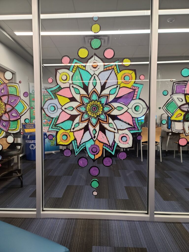  A brightly colored mandala with teal, yellow, pink, and purple shapes painted on a glass panel.
