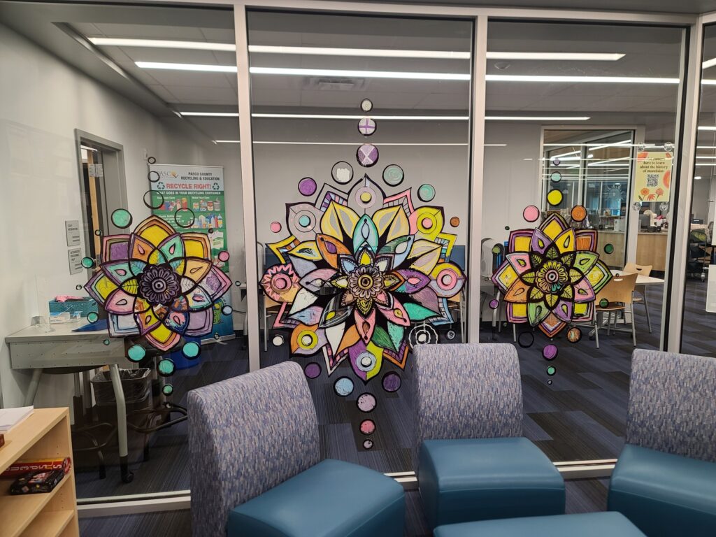 Three finished window mandalas displayed on large glass panels in the teen room, viewed from inside with blue seating in front.