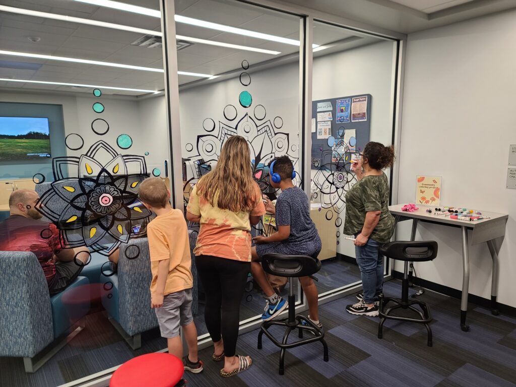 Children and adults coloring mandala outlines on the glass walls of a library room using chalk markers.