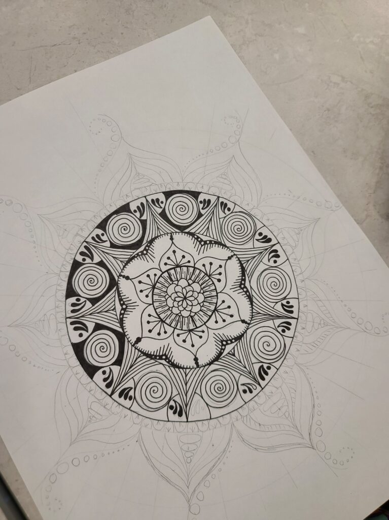 A highly detailed mandala drawn in black ink with swirling patterns, petals, and decorative linework on lightly sketched construction lines.
