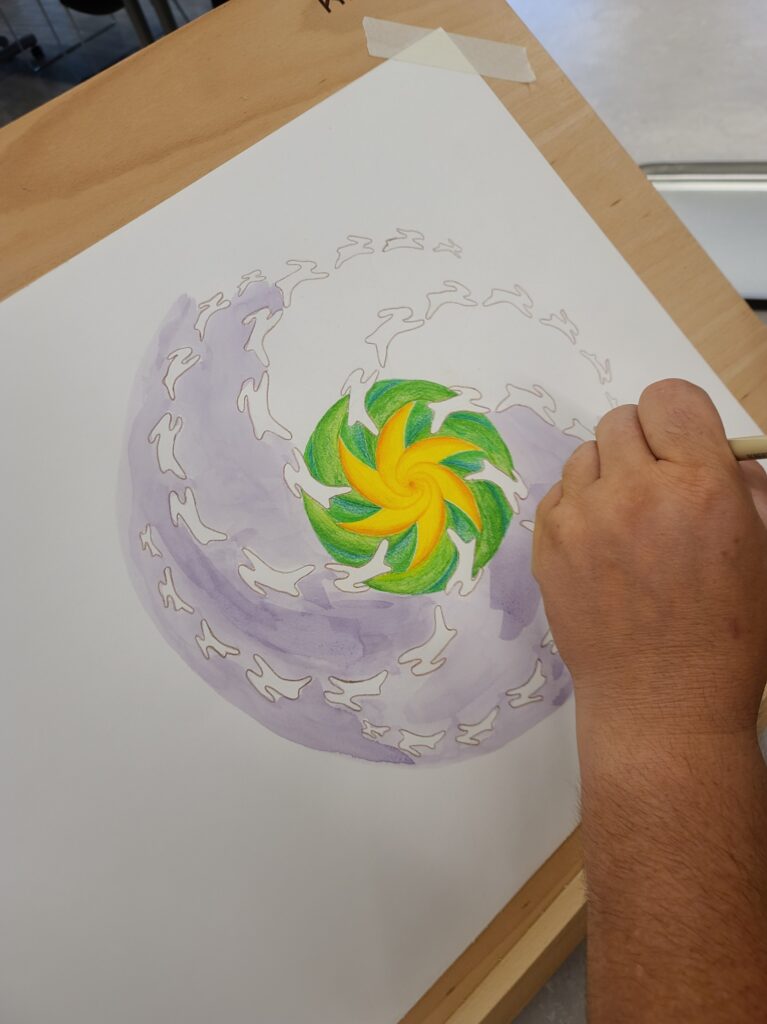 A close-up of a person’s hand drawing a spiral mandala with green and yellow shapes surrounded by pale purple watercolor rings.