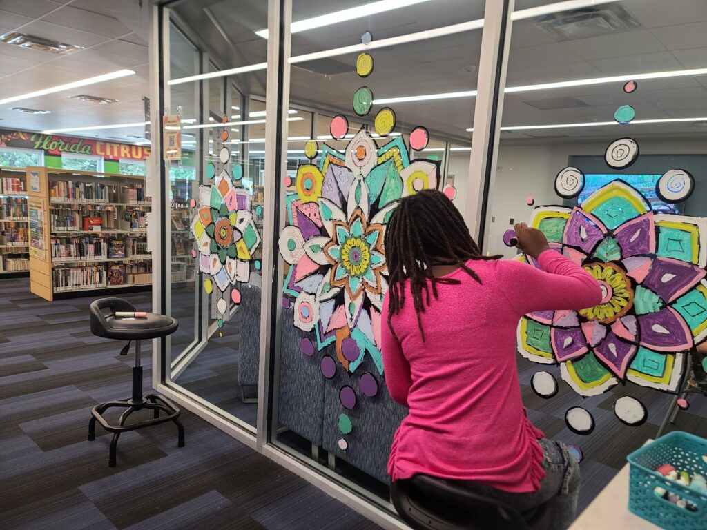  A teen wearing a bright pink shirt sits on a stool and colors a large mandala on the library window with chalk markers.
