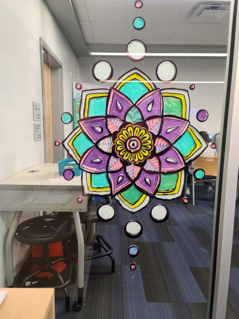  A colorful mandala with teal, purple, and yellow petals painted on a glass wall.
