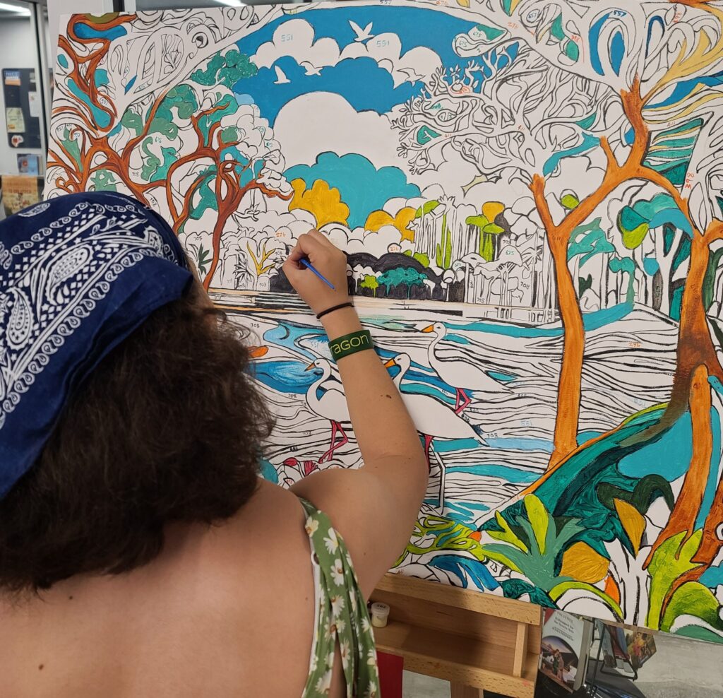 Back view of dark haired young woman in sun dress filing in a section of a large painting by hand.