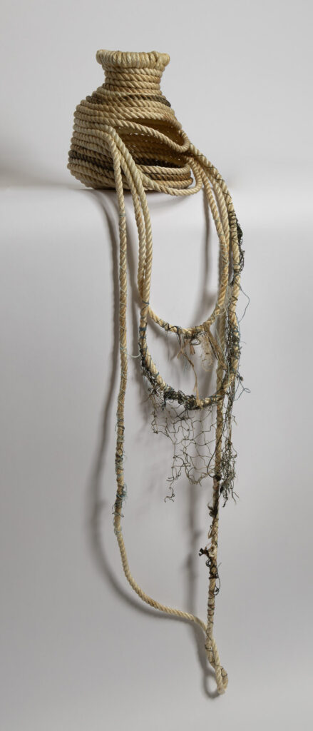 Sculptural vessel constructed from coiled cream and tan nylon rope, approximately basketball-sized, with a narrow neck at top. Long tailing strands of rope descend from the body, intertwined with deteriorating dark green ghost fishing net and marine debris. The piece leans against a neutral gray surface, casting a shadow that echoes it's form.