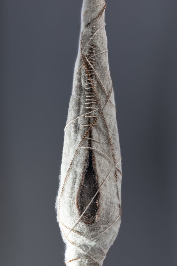 Close up of one felted wool pod form against grey wall, wrapped in thread, dark interior objects barely visible.