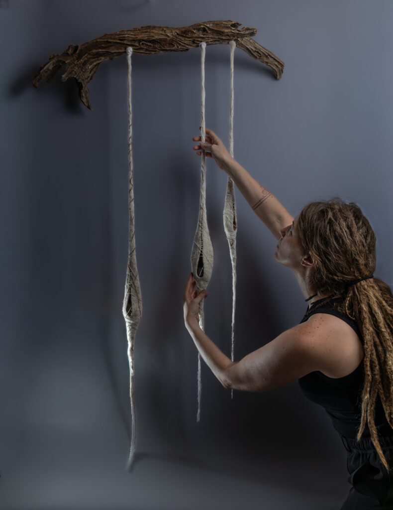 Artist in black clothing with long dreadlocks adjusts one of three felted pod shapes hanging from a piece of driftwood against a grey background. The pods are as big as her hand and hang down as long as her arm.