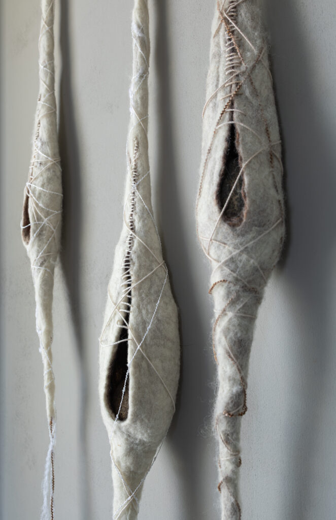 Three cream colored, long cocoon shapes hang against a warm gray background. Vertical slits reveal the dark interiors of the cocoon forms. Thread of various rough and soft textures are wrapped around the forms and small cream stitches partially close the openings.